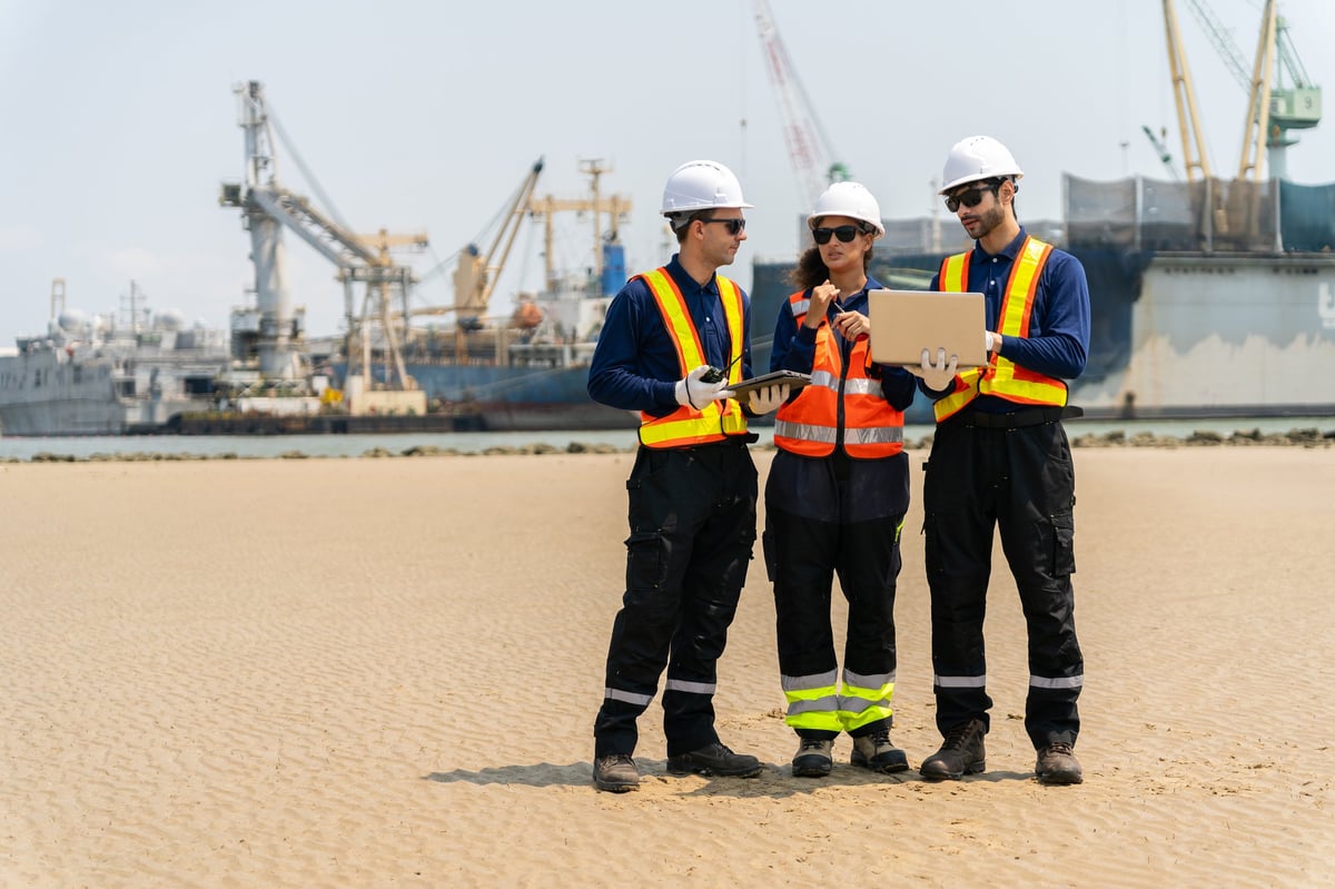 A group of engineers, wearing safety suits and helmets, are examining data on laptops in a shipyard and port area, with cranes and large ships in the background.