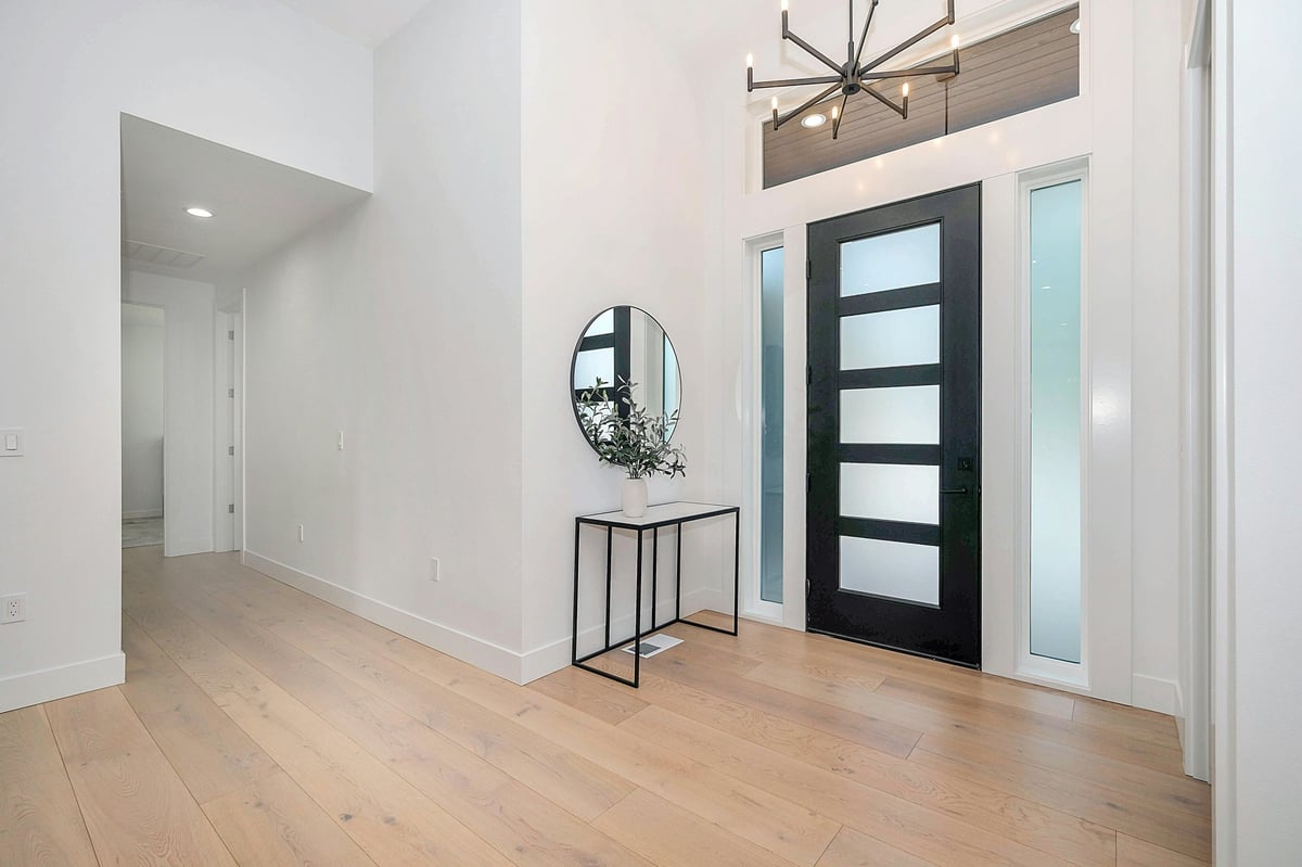 Modern foyer with light wood flooring, white walls, black front door with glass panels, round mirror, and metal console table