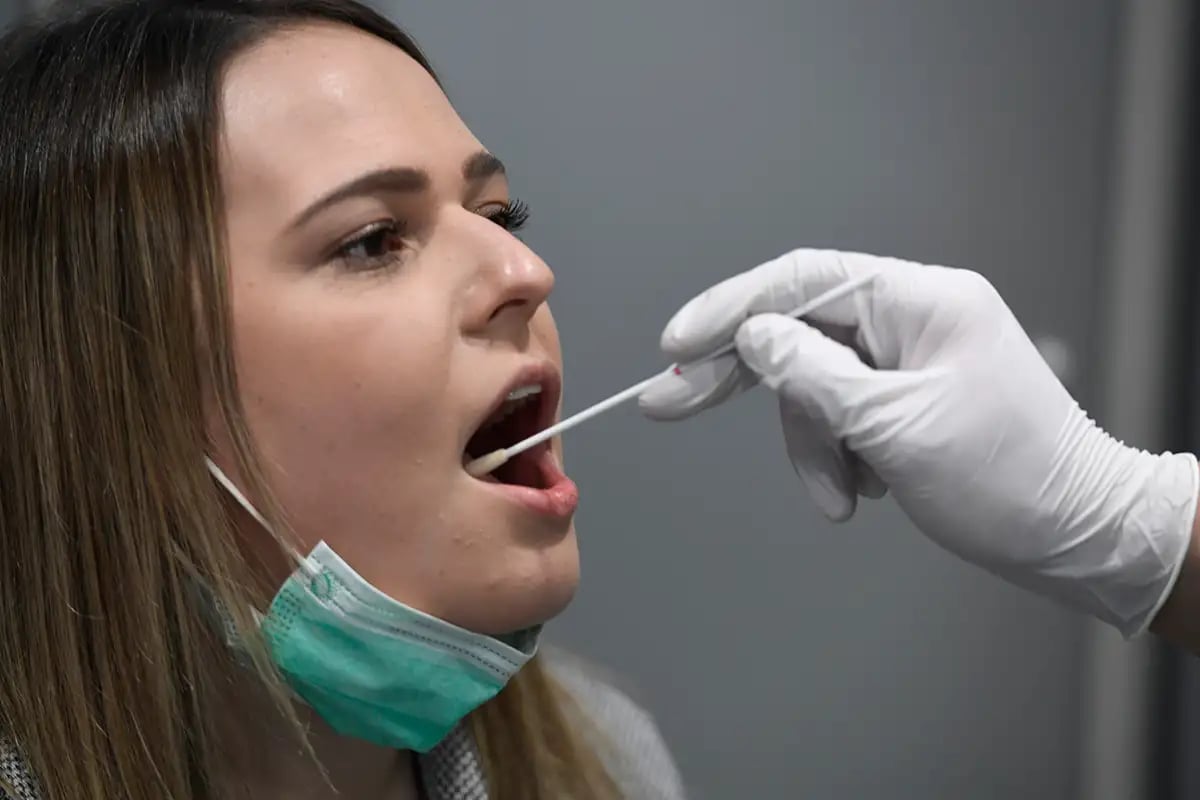 Woman receiving a COVID-19 nasal swab test from a healthcare worker wearing white gloves, with a protective mask around her neck.