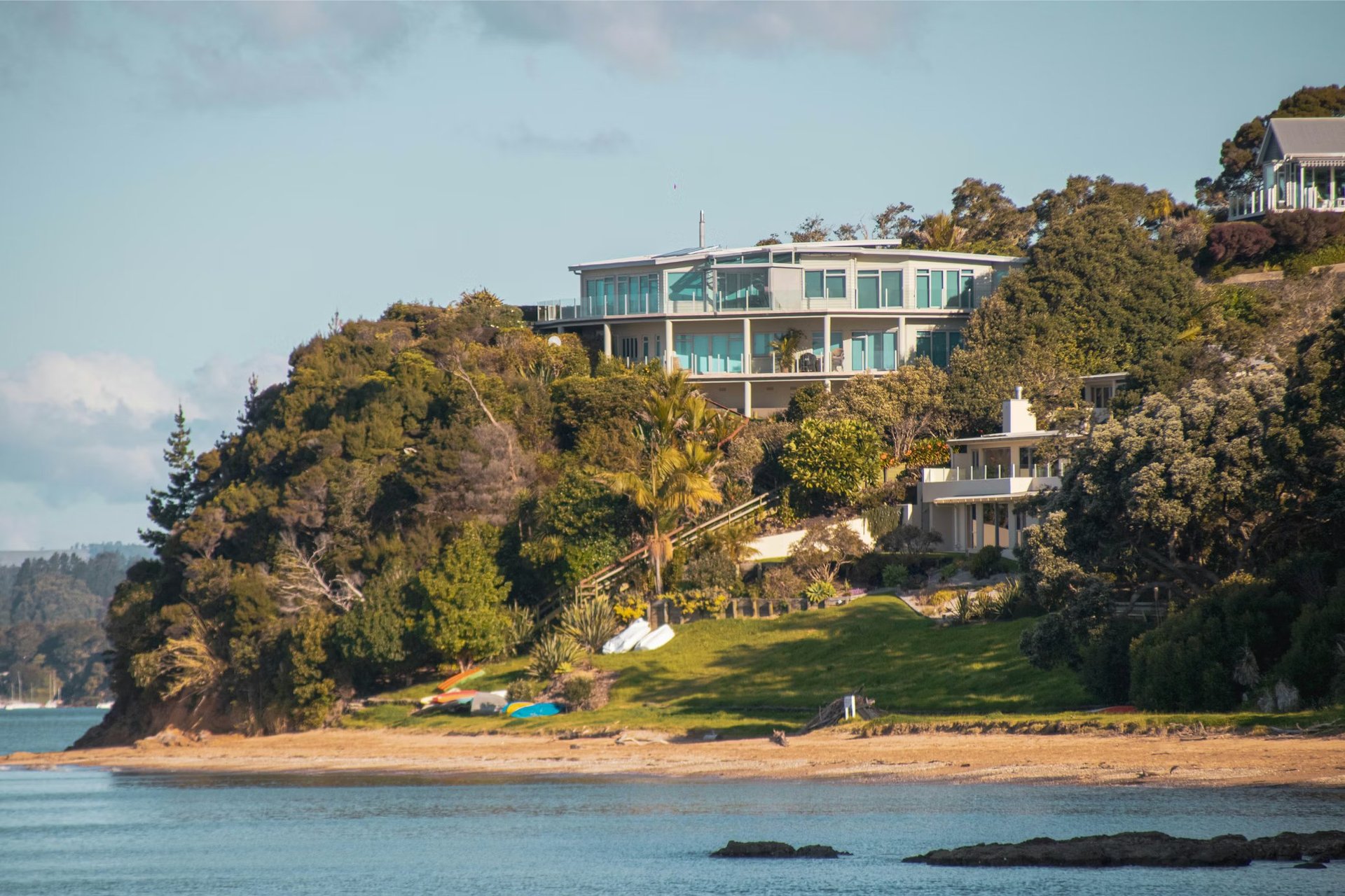 Modern multi-story beachfront mansion on a forested hillside overlooking calm blue water with a sandy beach below.